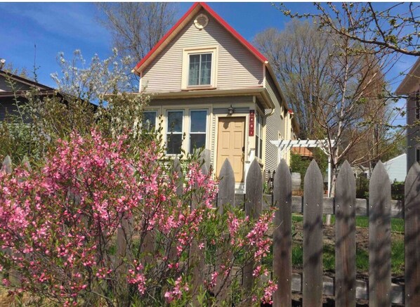Una casa de dos pisos con techo rojo y revestimiento color beige, un porche delantero y una cerca de piquetes de madera.