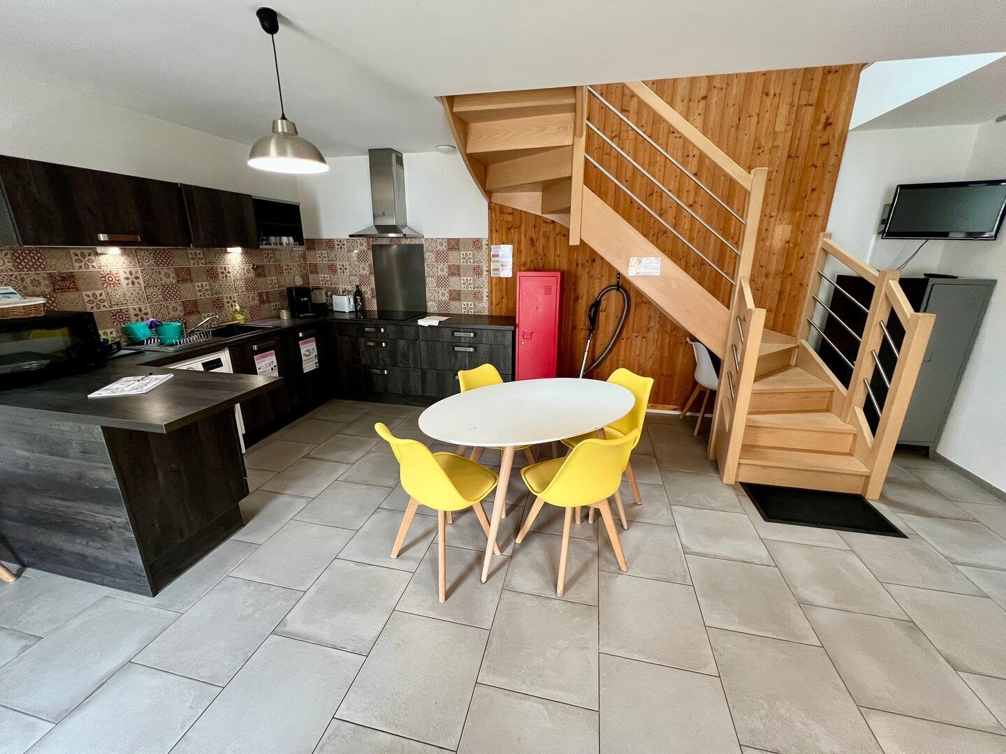A modern kitchen with a central island, yellow chairs, a wooden staircase, and a TV mounted on the wall.