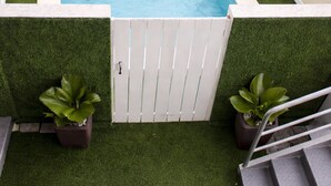 Una piscina rectangular con borde blanco, rodeada de césped artificial. Se observa una reja blanca, dos plantas en macetas y un banco pequeño.