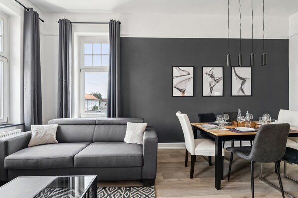 A modern living room with a grey sofa, a dining table set for four, and abstract art on the wall. - Charysma: Jewel in anthracite | elegant | stylish | 5 min walk to the old town (Hameln)