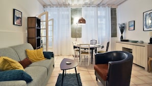 Una sala de estar con sofá, una mesa de centro, una mesa de comedor con sillas, una lámpara y un área de cocina con alacena y un florero.