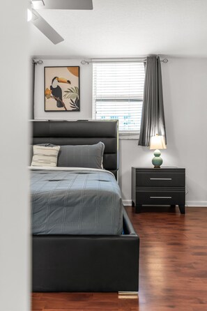 A bedroom with a bed, a nightstand, a lamp, a window with blinds, and a wall-mounted picture. - Perfectly Located Miami 3BR with Stunning Views (Miami)