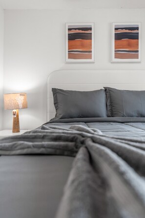 A neatly made bed with grey bedding, a wooden bedside table with a lamp, and two framed abstract paintings on the wall.