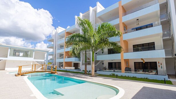 Un edifici modern de diversos pisos amb una piscina, palmeres i un cel blau clar.