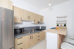 A modern kitchen with wooden cabinets, a stainless steel refrigerator, a microwave, a sink, and a countertop with a cutting board and fruits.
