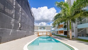 Una piscina con cubierta de madera, una palmera y edificios modernos al fondo.