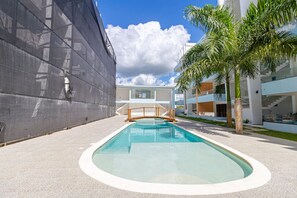 Une piscine avec une terrasse en bois, un palmier et des bâtiments modernes en arrière-plan.