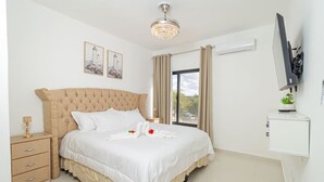 Un dormitorio con una cama grande, una mesita de noche, un televisor montado en la pared y una ventana con cortinas.