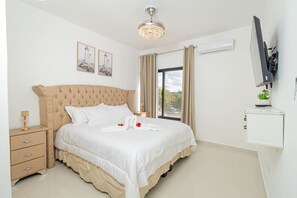 Een slaapkamer met een groot bed, een nachtkastje, een aan de muur bevestigde televisie en een raam met gordijnen.