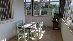 Quarto iluminado pelo sol com soalho em mosaico, uma mesa com cadeiras, uma janela com vista para árvores e edifícios, e um vaso de planta.