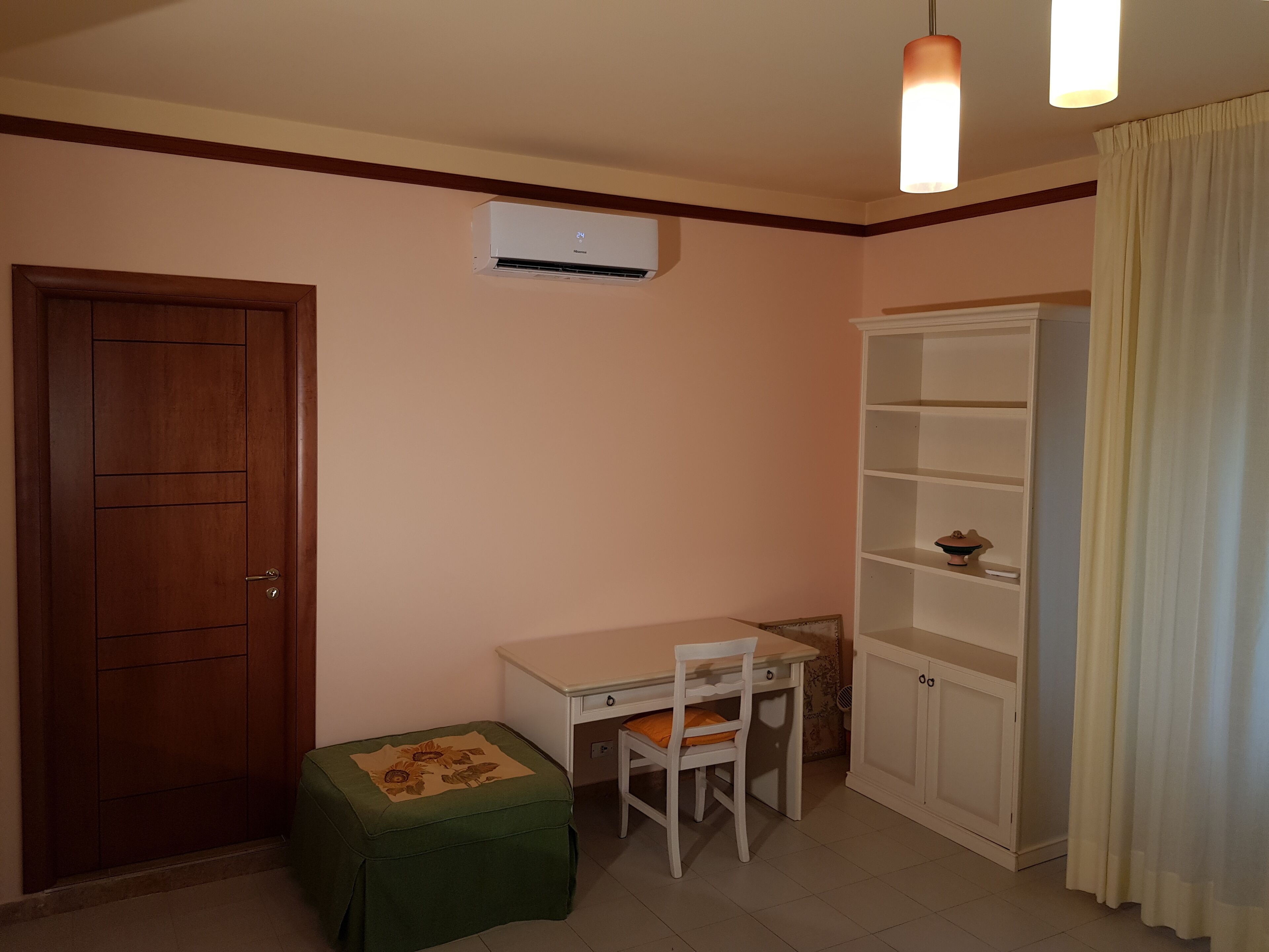 A small room with a wooden door, a white desk with a chair, a green ottoman, a white cabinet, and a ceiling-mounted air conditioner.