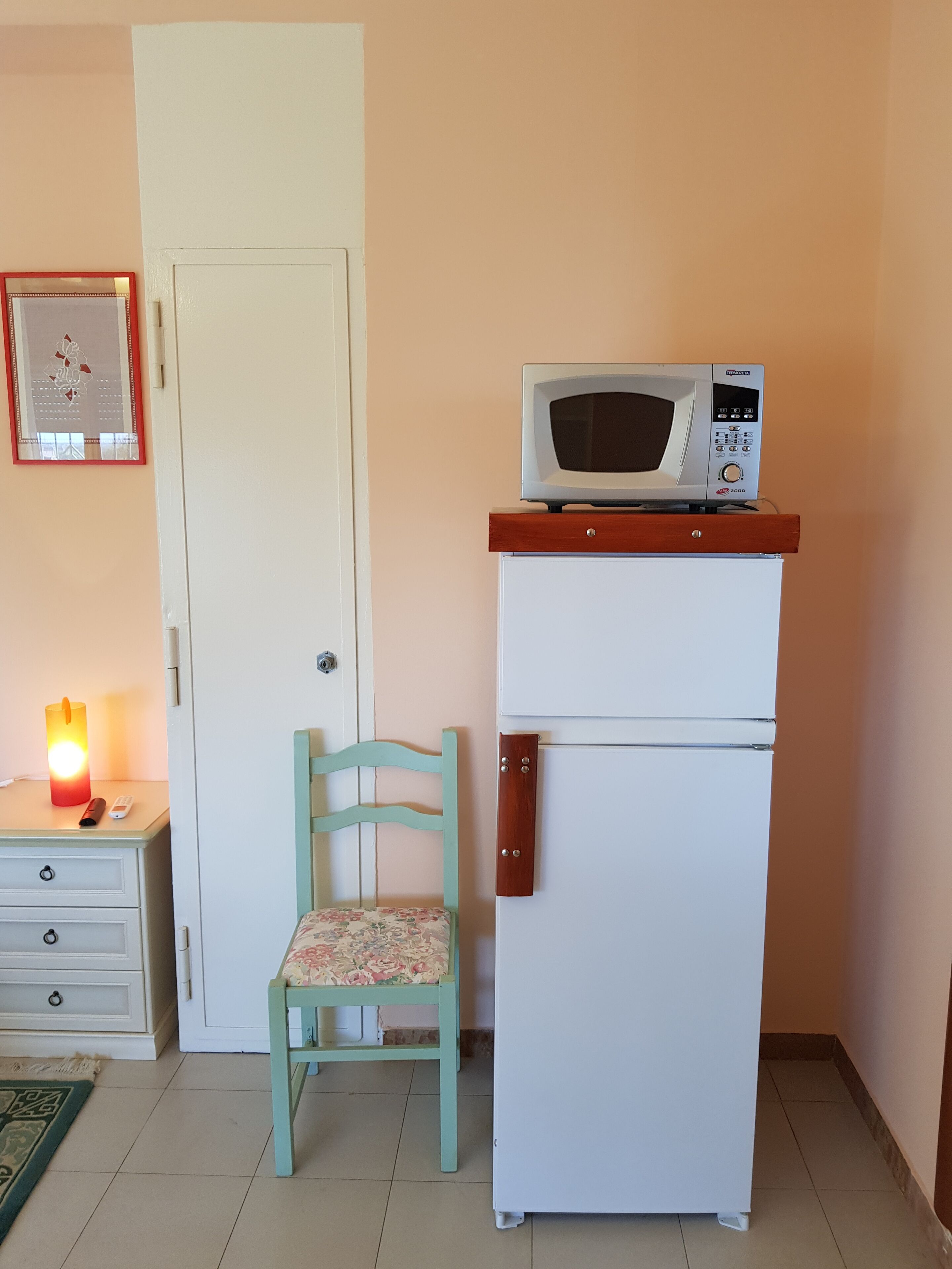 Eine kompakte Küchenzeile mit einer Mikrowelle auf einem Kühlschrank, ein Holzsessel mit einem floralen Kissen, ein Nachttisch mit Fernbedienung und Kerze sowie eine Tür mit einem dekorativen Spiegel.