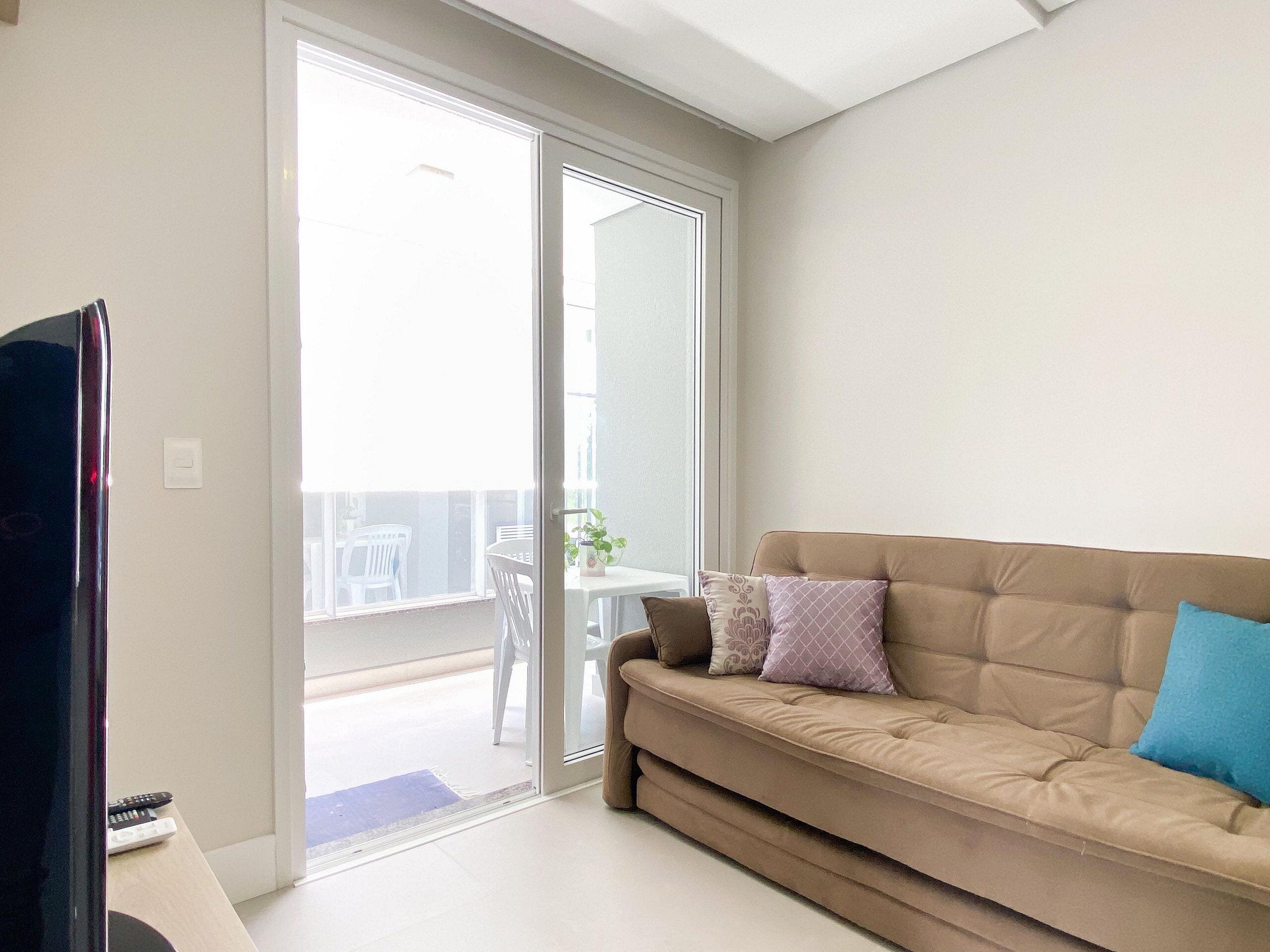 Una sala de estar con un sofá color beige, un televisor y un ventanal corredero que da a un espacio exterior con una mesa y sillas.