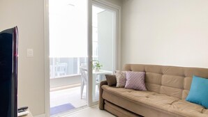 A living room with a beige sofa, a television, and a sliding glass door leading to an outdoor area with a table and chairs. - Apê Cheio de Estilo - Campeche (Florianopolis)