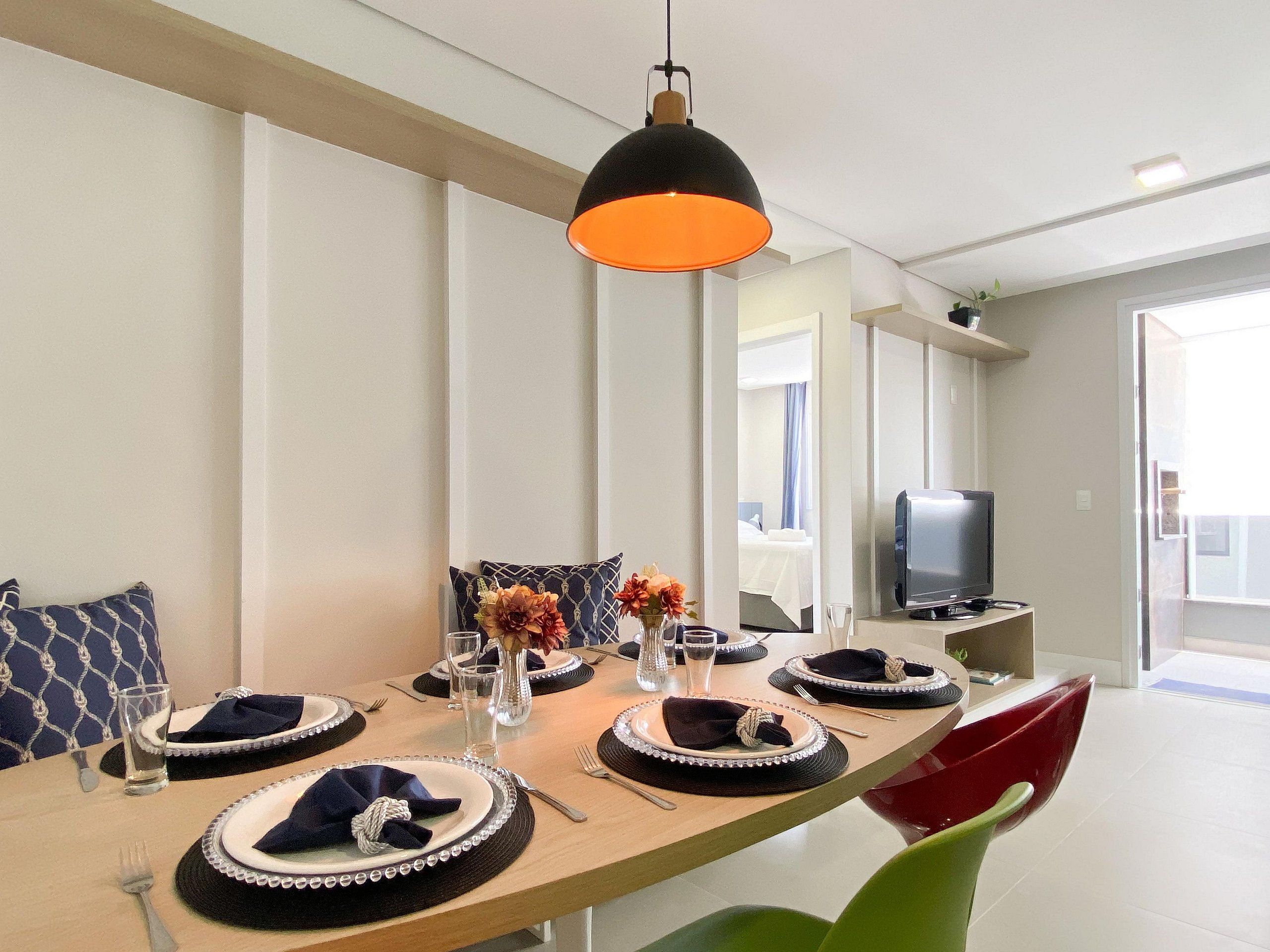 Un coin repas avec une longue table dressée pour quatre personnes, un téléviseur posé sur un meuble et un luminaire suspendu au design moderne.
