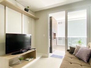 A living room with a flat-screen TV on a wooden stand, a sofa with cushions, a small plant on a shelf, and a sliding glass door leading to a balcony with outdoor furniture. - Apê Cheio de Estilo - Campeche (Florianopolis)