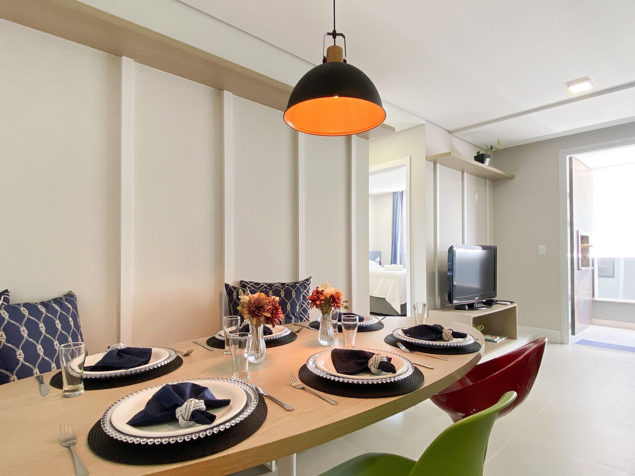 Un comedor con una mesa larga dispuesta para cuatro personas, un televisor sobre un mueble bajo y una lámpara colgante moderna.