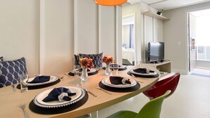 A dining area with a long table set for four, a television on a stand, and a modern pendant light. - Apê Cheio de Estilo - Campeche (Florianopolis)