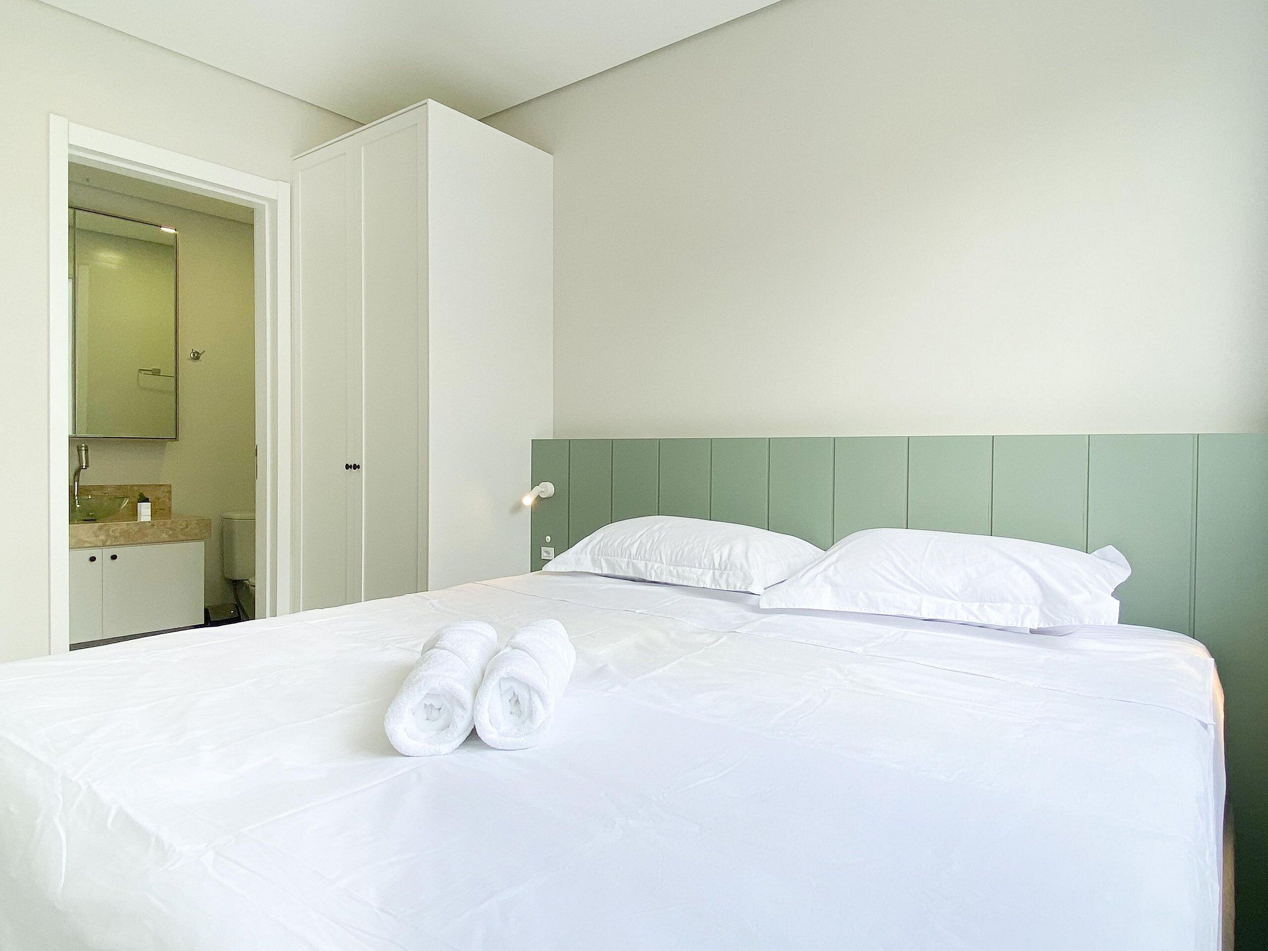 Una cama bien hecha con sábanas blancas y dos toallas enrolladas, un cabecero verde y un armario blanco con espejo.