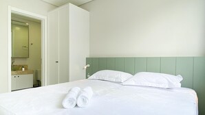 A neatly made bed with white linens and two rolled towels, a green headboard, and a white wardrobe with a mirror. - Apê Cheio de Estilo - Campeche (Florianopolis)