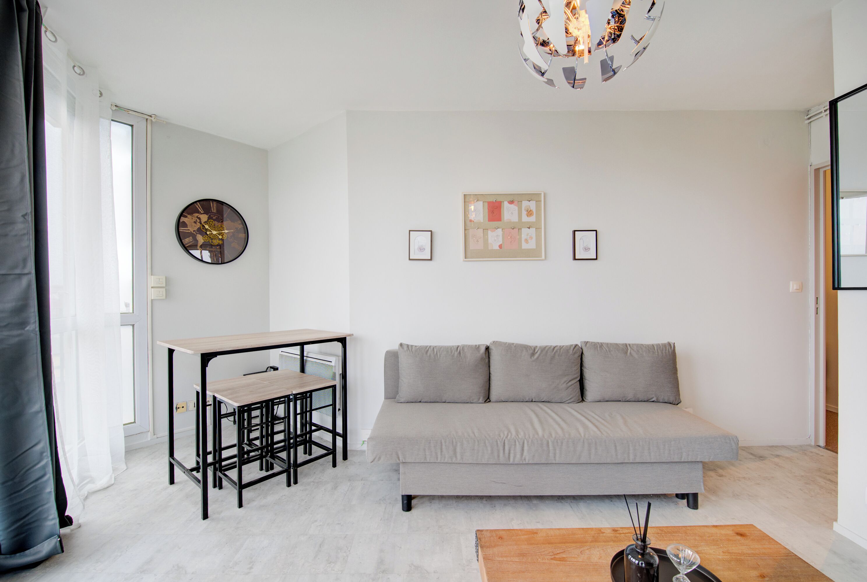 Uma sala de estar moderna com um sofá cinzento, uma mesa de madeira pequena com duas cadeiras, um relógio de parede e quadros emoldurados.