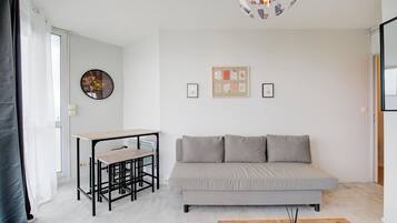 Uma sala de estar moderna com um sofá cinzento, uma mesa de madeira pequena com duas cadeiras, um relógio de parede e quadros emoldurados.