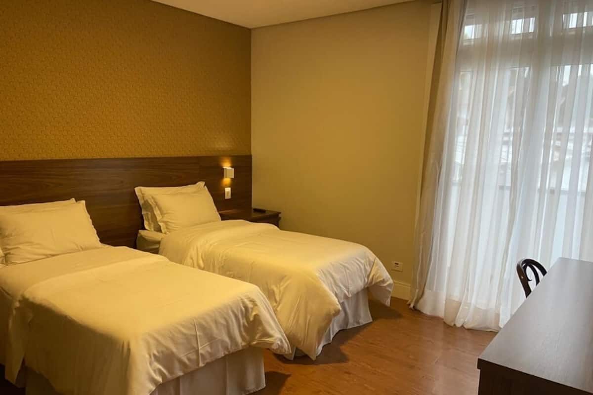 Une chambre d’hôtel avec deux lits simples, une tête de lit en bois, une table de chevet, un bureau et une fenêtre avec des rideaux légers.