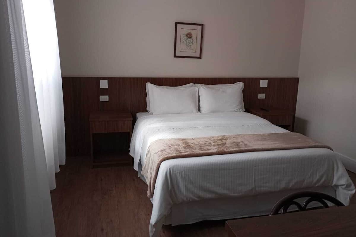Une chambre d’hôtel avec un lit, une table de chevet, une tête de lit en bois, un tableau encadré au mur et une fenêtre avec des rideaux.