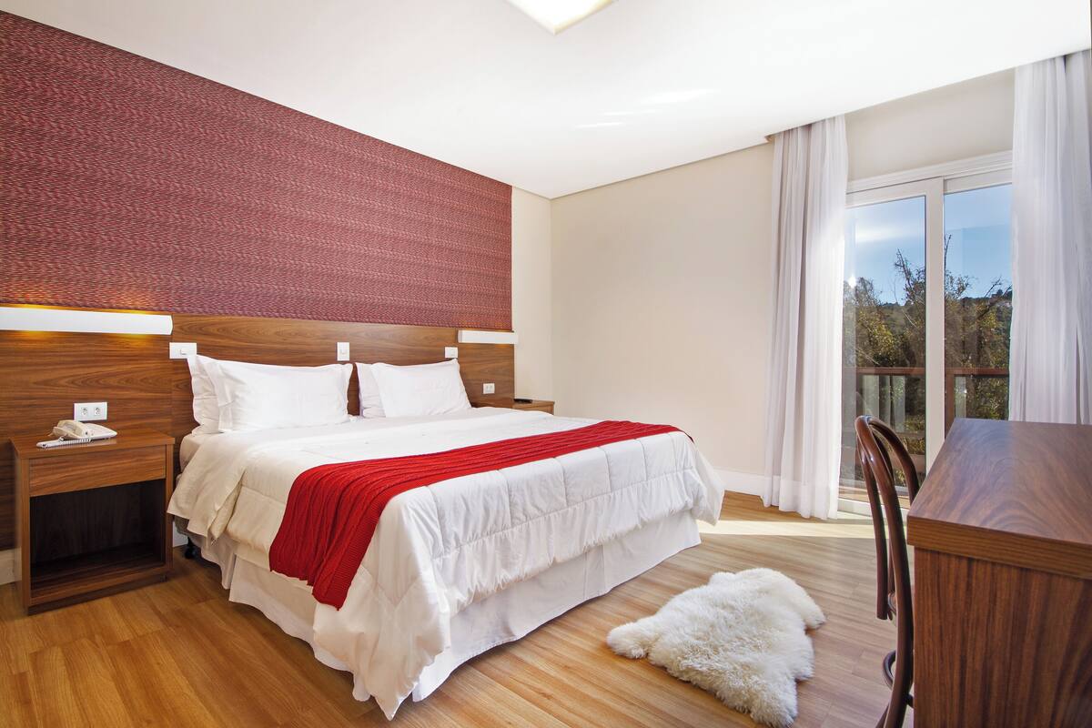 Une chambre moderne avec un grand lit, une tête de lit en bois, des tables de chevet et une vue sur l’extérieur grâce à une grande fenêtre.