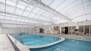 An indoor swimming pool with a glass ceiling, white tiled walls, and a seating area with tables and chairs.