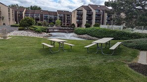 Un patio con mesas de picnic, una pequeña fuente de agua y un edificio con balcones al fondo.