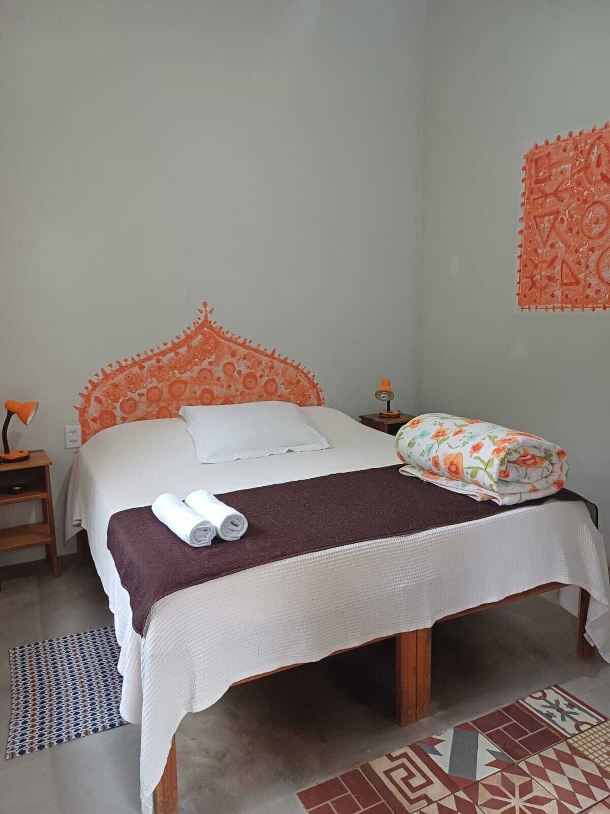 A bed with a white and orange headboard, a white bedspread, and a brown blanket. There are two pillows, one white and one with a floral pattern. A pair of rolled towels is on the bed.