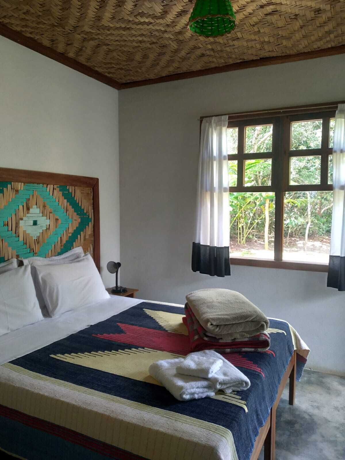 A bedroom with a bed, a window with curtains, a woven ceiling, and a wall with a geometric pattern.