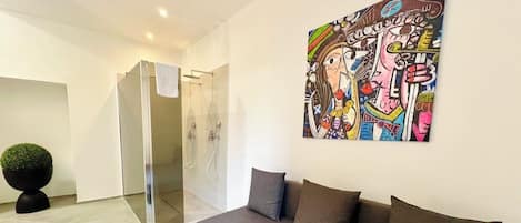 A modern interior with a glass-enclosed shower, a grey sofa with cushions, and a colorful abstract painting on the wall.