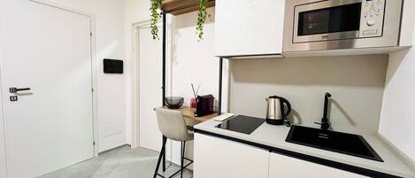 A compact kitchen with white cabinets, a stainless steel microwave, a kettle, and a small bar stool.
