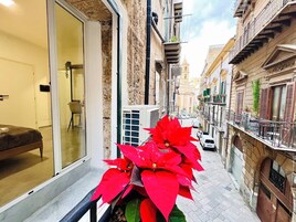 Una calle estrecha con un balcón, una planta en maceta y un edificio que muestra una puerta y una ventana.