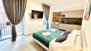 A modern hotel room with a bed, a TV, a kitchenette, and a balcony with curtains. - PRETORIA SUITES (Palermo)