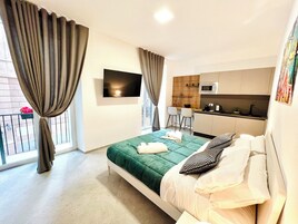 Una habitación de hotel moderna con cama, televisión, kitchenette y balcón con cortinas.