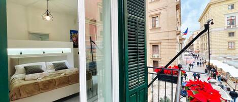 Quarto de hotel com cama, luminária pendente, varanda com porta verde e vista para a rua.