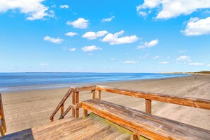 Un paseo marítimo de madera que conduce a una playa arenosa bajo un cielo azul despejado con nubes dispersas.