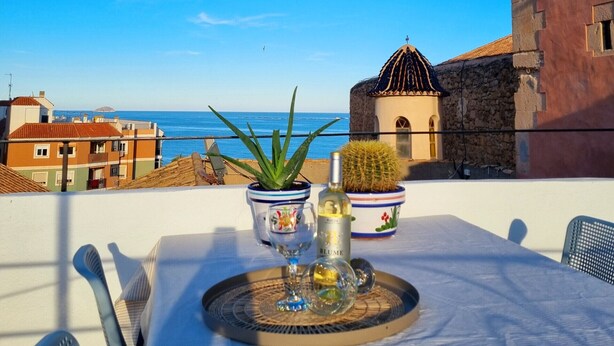 A table with a clock, potted plants, and a bottle on a balcony overlooking a coastal view.