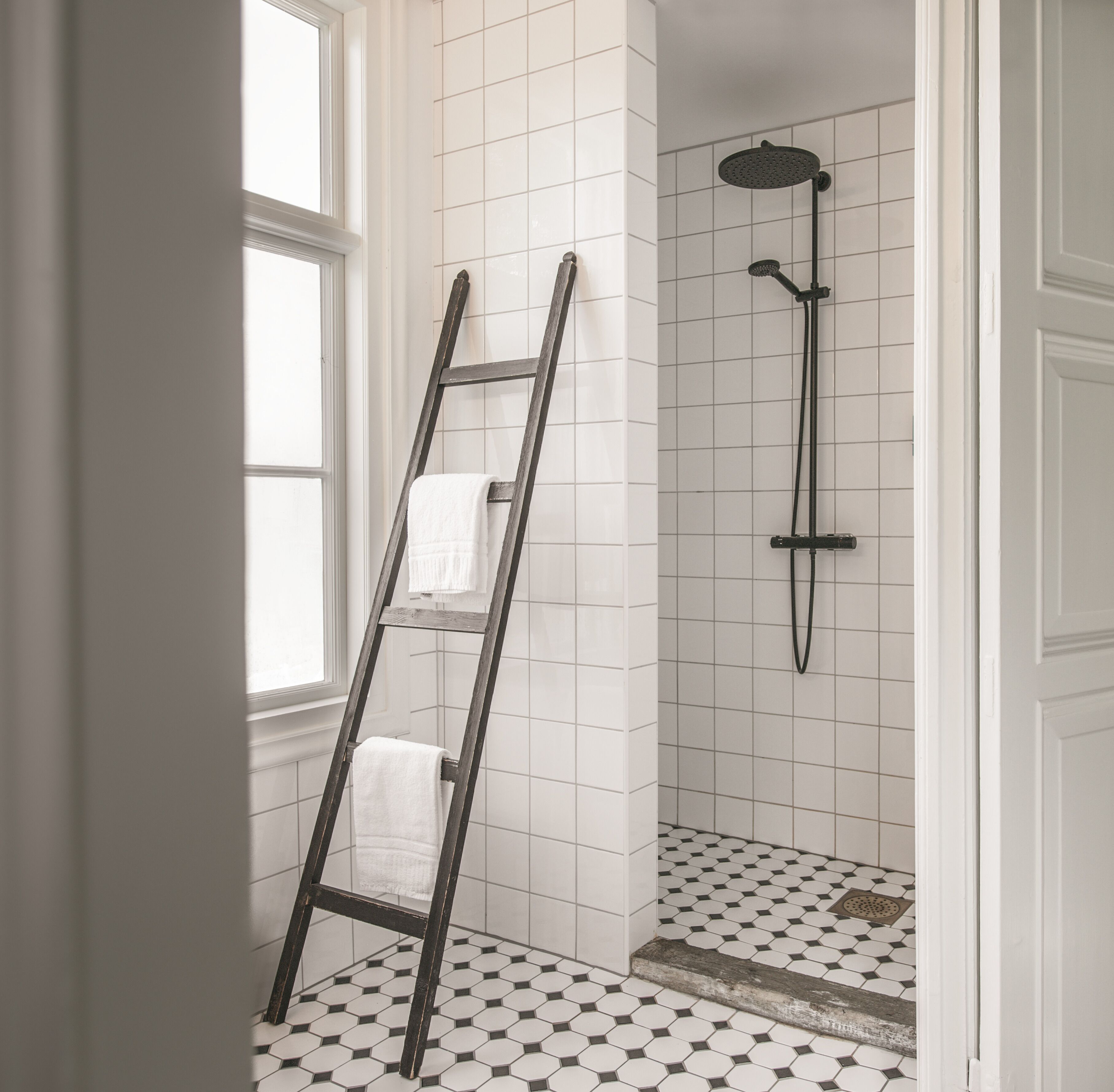 Un baño con paredes de azulejos blancos, una ducha con rociador negro y una escalera con toallas.