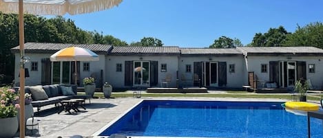 A swimming pool with a patio area, outdoor furniture, and a building with multiple doors and windows.