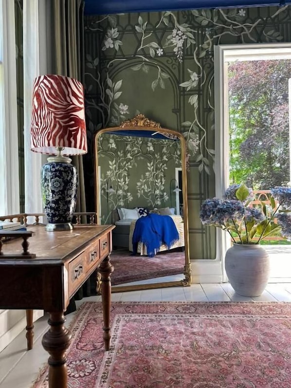 A bedroom with a bed, a mirror, a lamp, a table, and a large window.