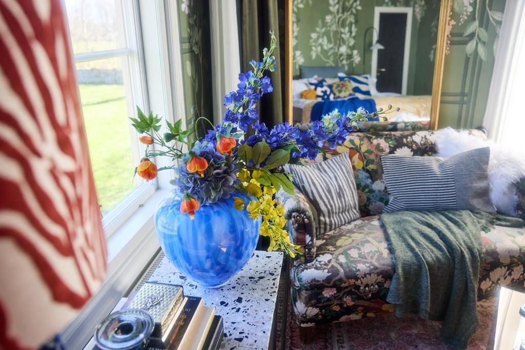 A room with a large window, a blue vase with flowers, a floral sofa, and a mirror reflecting the room.