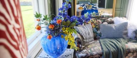 A room with a large window, a blue vase with flowers, a floral sofa, and a mirror reflecting the room.