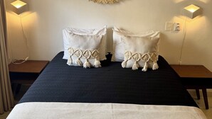 Una cama con cabecera oscura, dos almohadas blancas con flecos y un tapiz tejido en la pared.