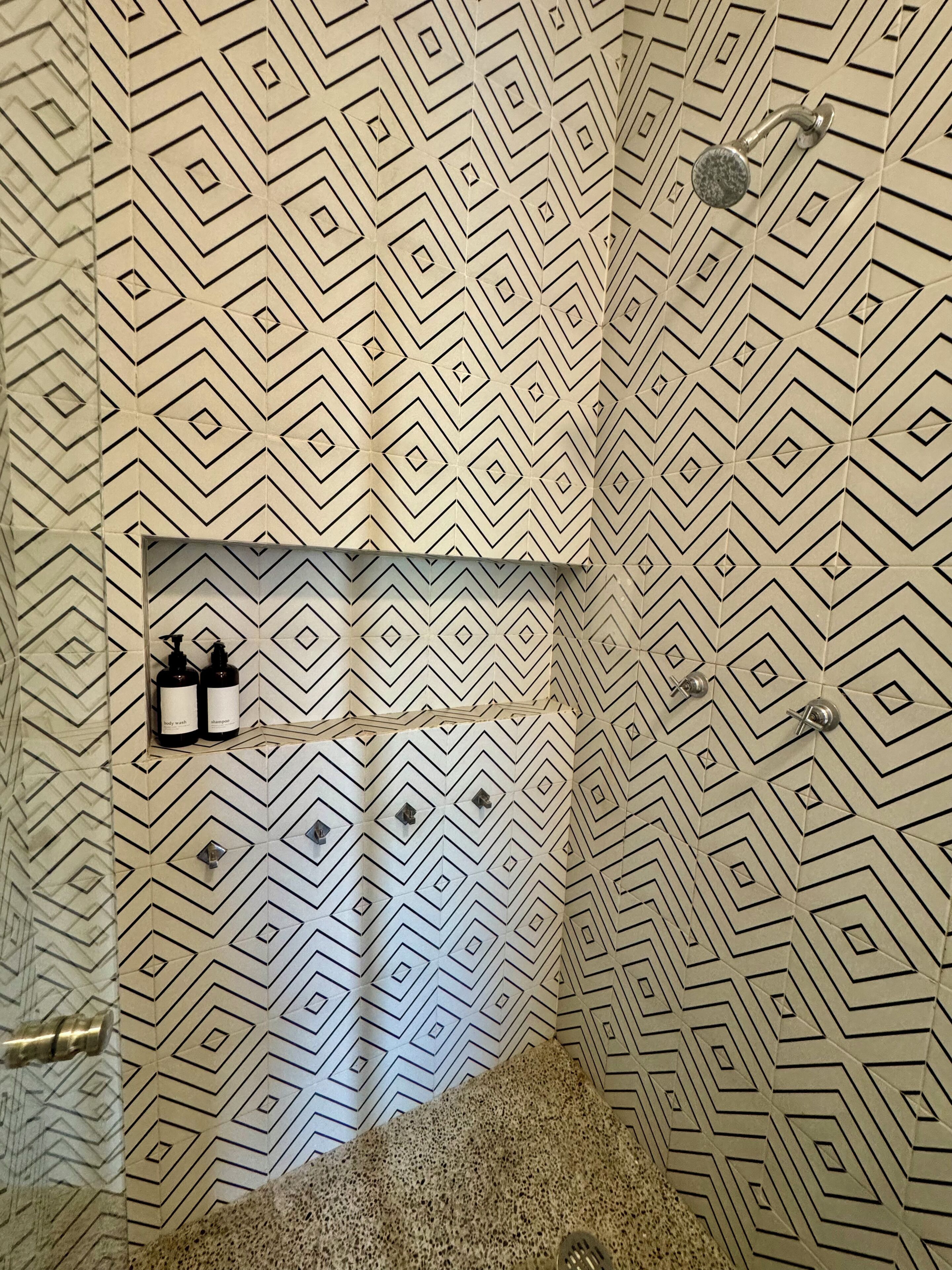Un baño con un diseño de azulejos geométrico, zona de ducha y estante con dos botellas que dicen "body wash" y "champú".