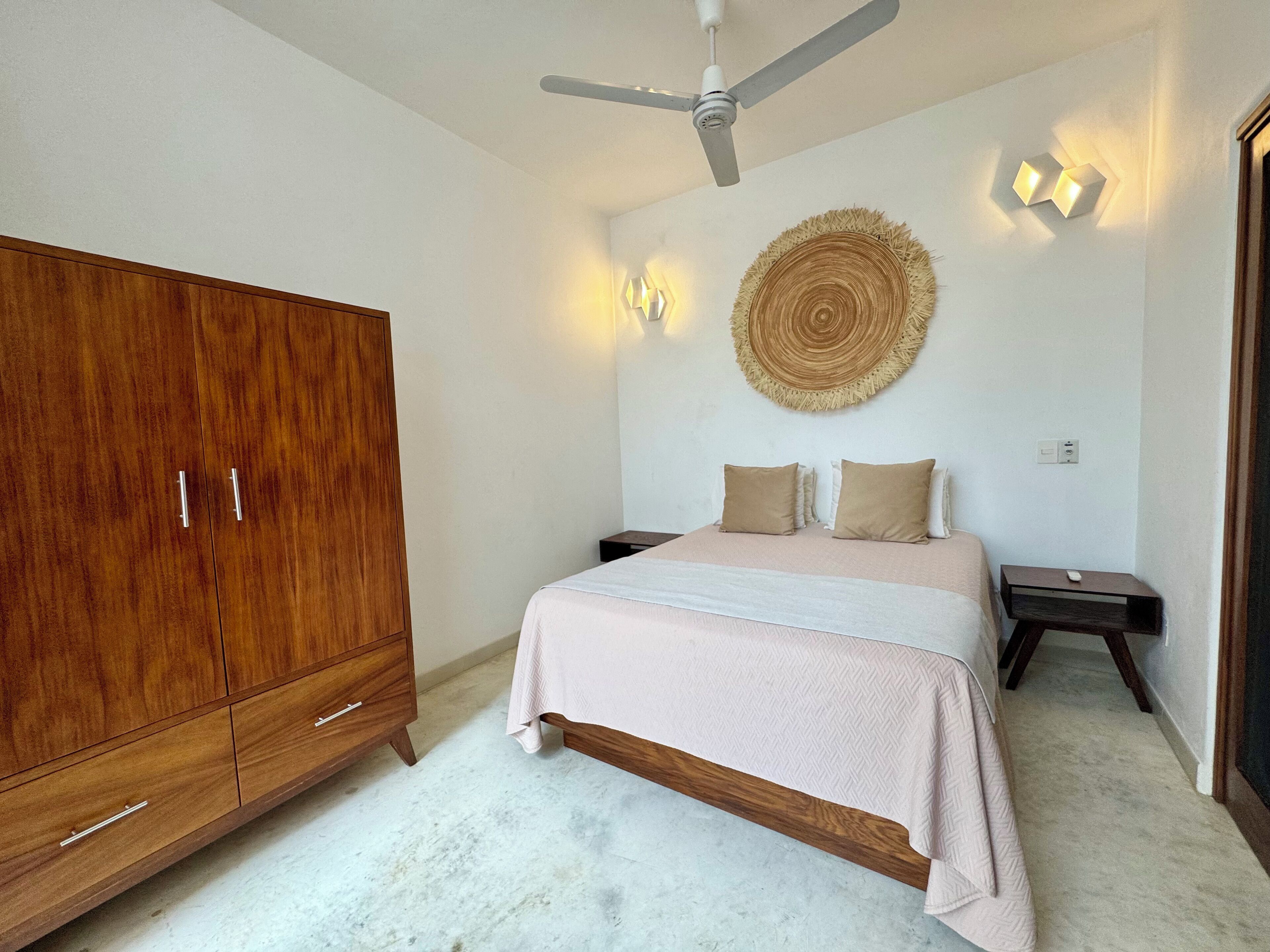 A bedroom with a large wooden wardrobe, a bed with a white and beige bedspread, two bedside tables with lamps, and a ceiling fan.