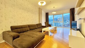 Ein modernes Wohnzimmer mit einer Eckcouch, einem kleinen weißen Tisch und einem großen Fenster mit Blick auf die Stadt.